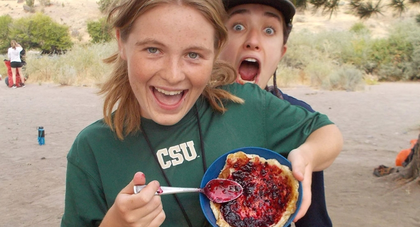 A student holds up a fruity dish and smiles at the camera, while another person smiles behind them. 
